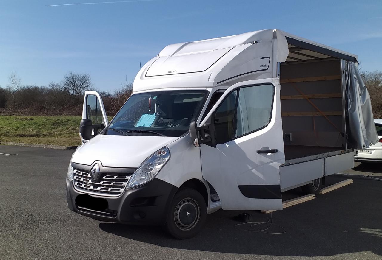 Biały Renault Master z otwartymi drzwiami kabiny i przestrzeni ładunkowej, zaparkowany na parkingu z widocznym fragmentem innego pojazdu w tle.