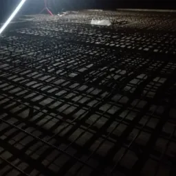 Siatka zbrojeniowa ułożona na betonowej powierzchni, oświetlona przez mocne światło lampy budowlanej, widoczna w tle, tworząca geometryczny wzór cieni.