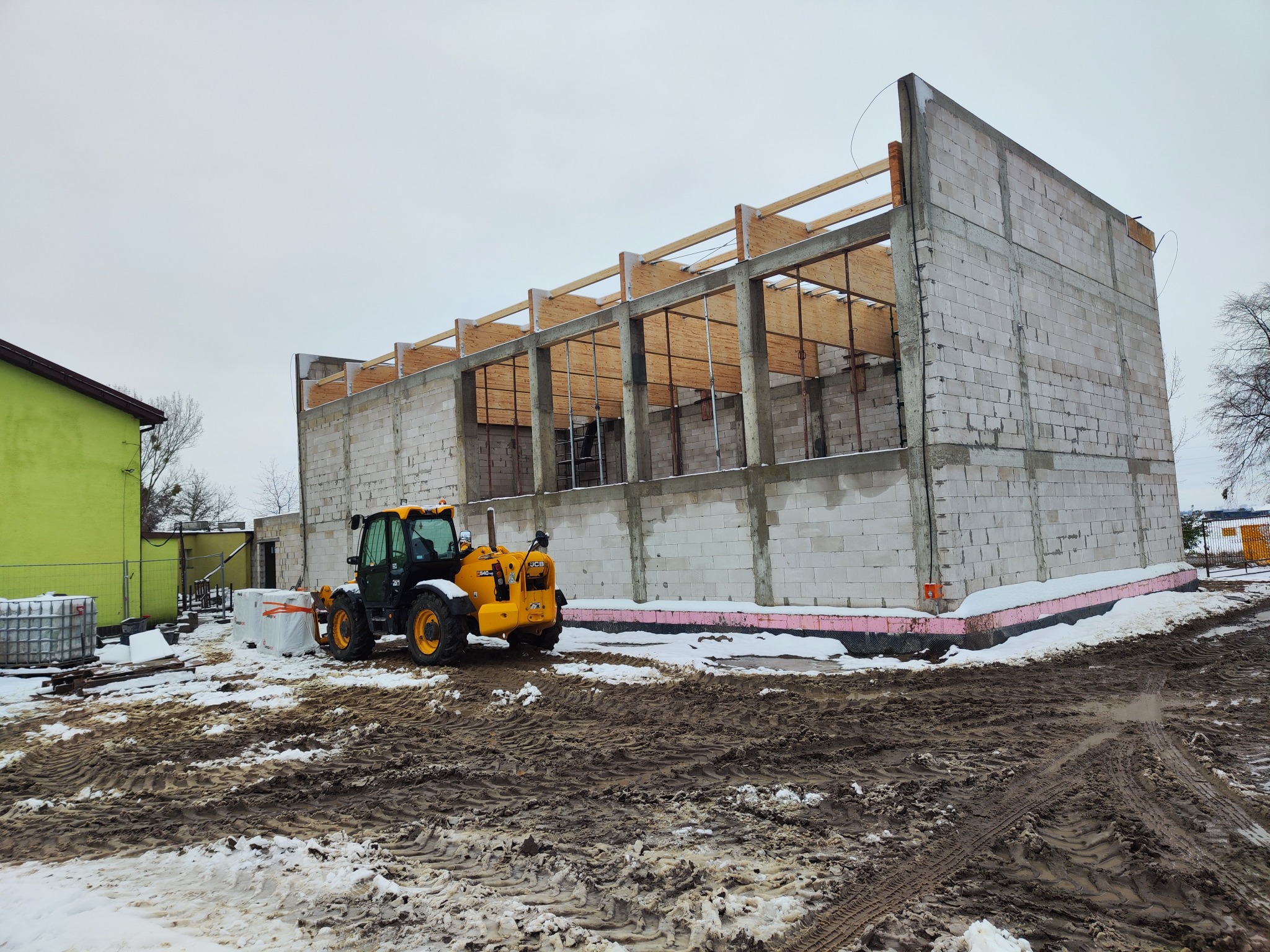 Budowa hali z bloczków betonu komórkowego w zimowej scenerii. Żółta ładowarka teleskopowa JCB na pierwszym planie. Widoczne drewniane elementy konstrukcji dachu.