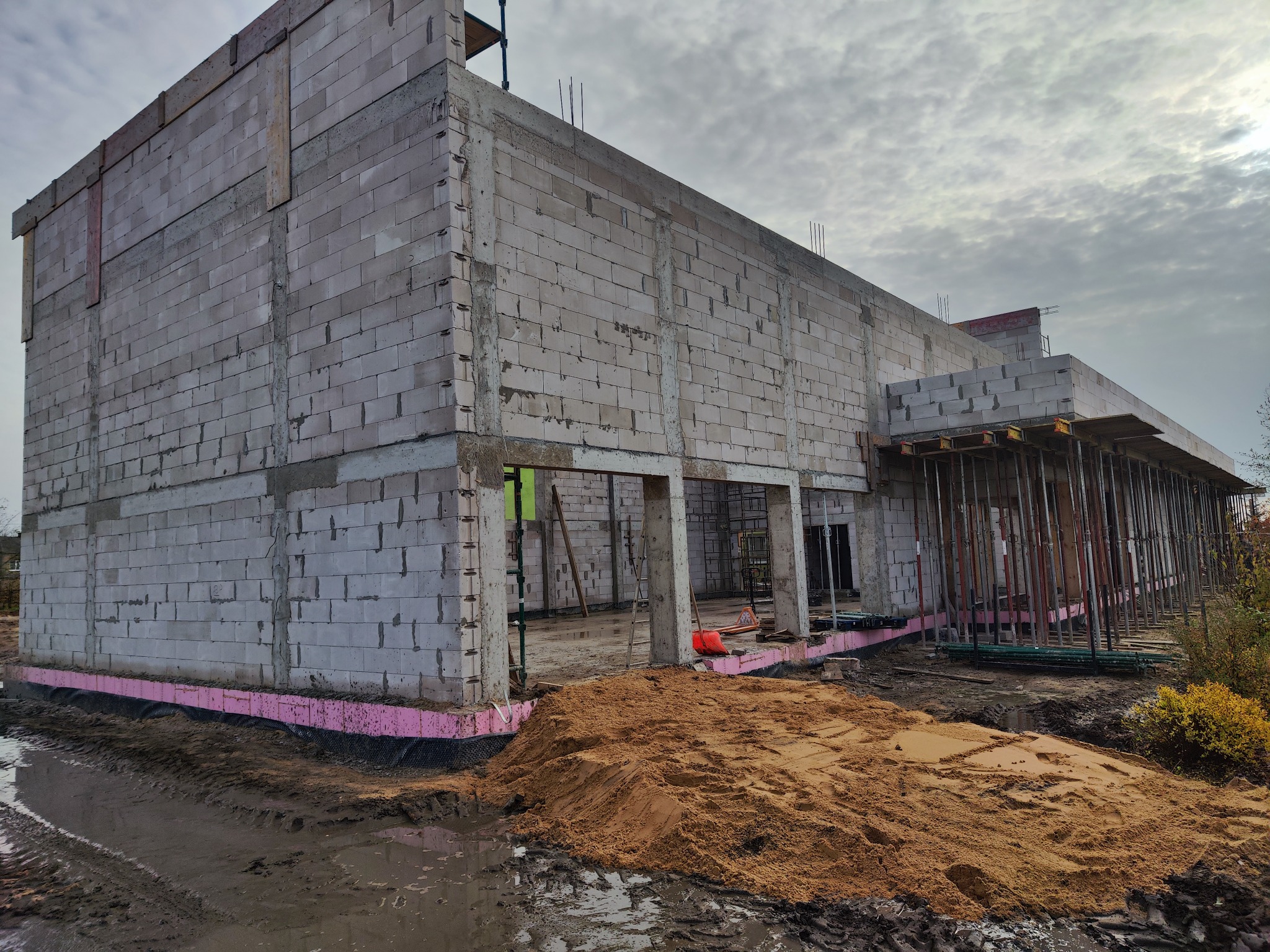 Surowy stan domu z białego bloczka, widoczne szalunki i izolacja fundamentów w kolorze różowym. Na pierwszym planie piasek i błoto. Pochmurne niebo w tle.