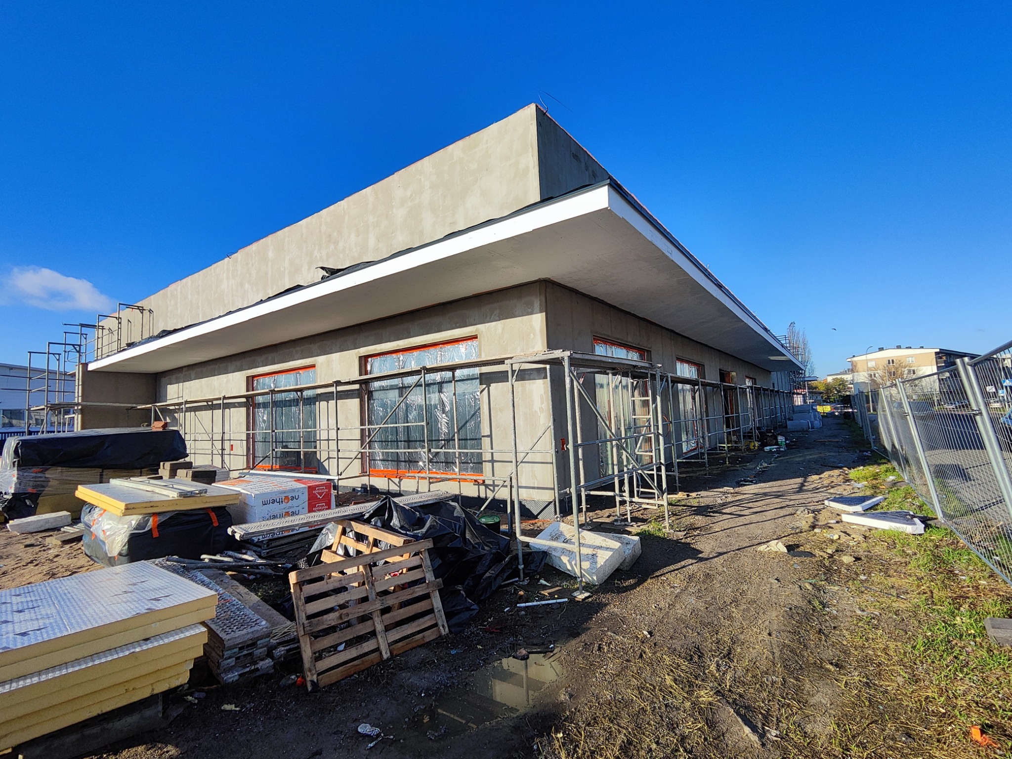 Budynek w trakcie budowy z szarymi ścianami, rusztowaniami i materiałami budowlanymi na ziemi, pod błękitnym niebem. Widoczne okna zabezpieczone folią.