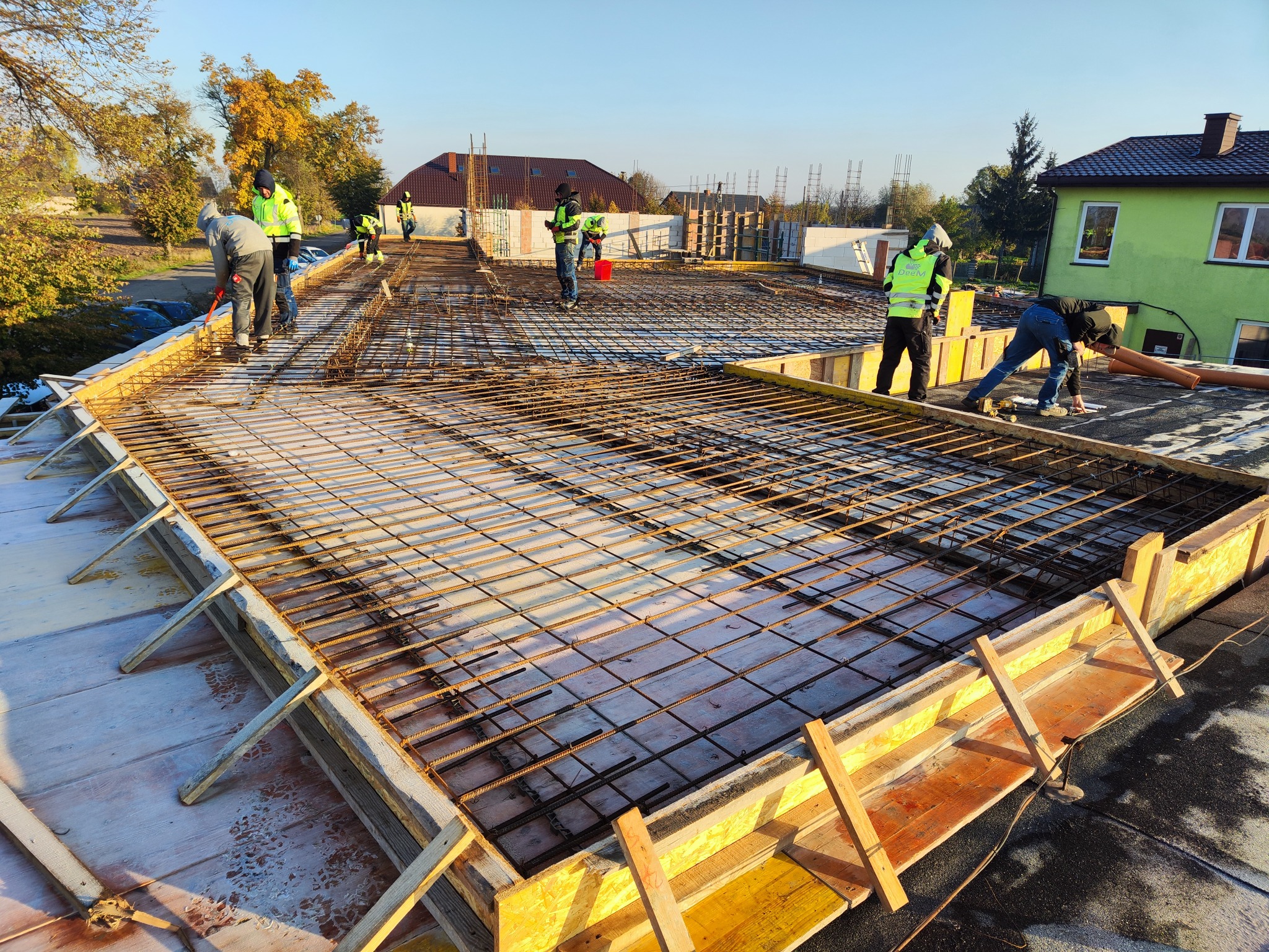 Zbrojenie płyty fundamentowej z drewnianym szalunkiem, widoczni pracownicy w odblaskowych kamizelkach, w tle budynki w trakcie budowy i drzewa.