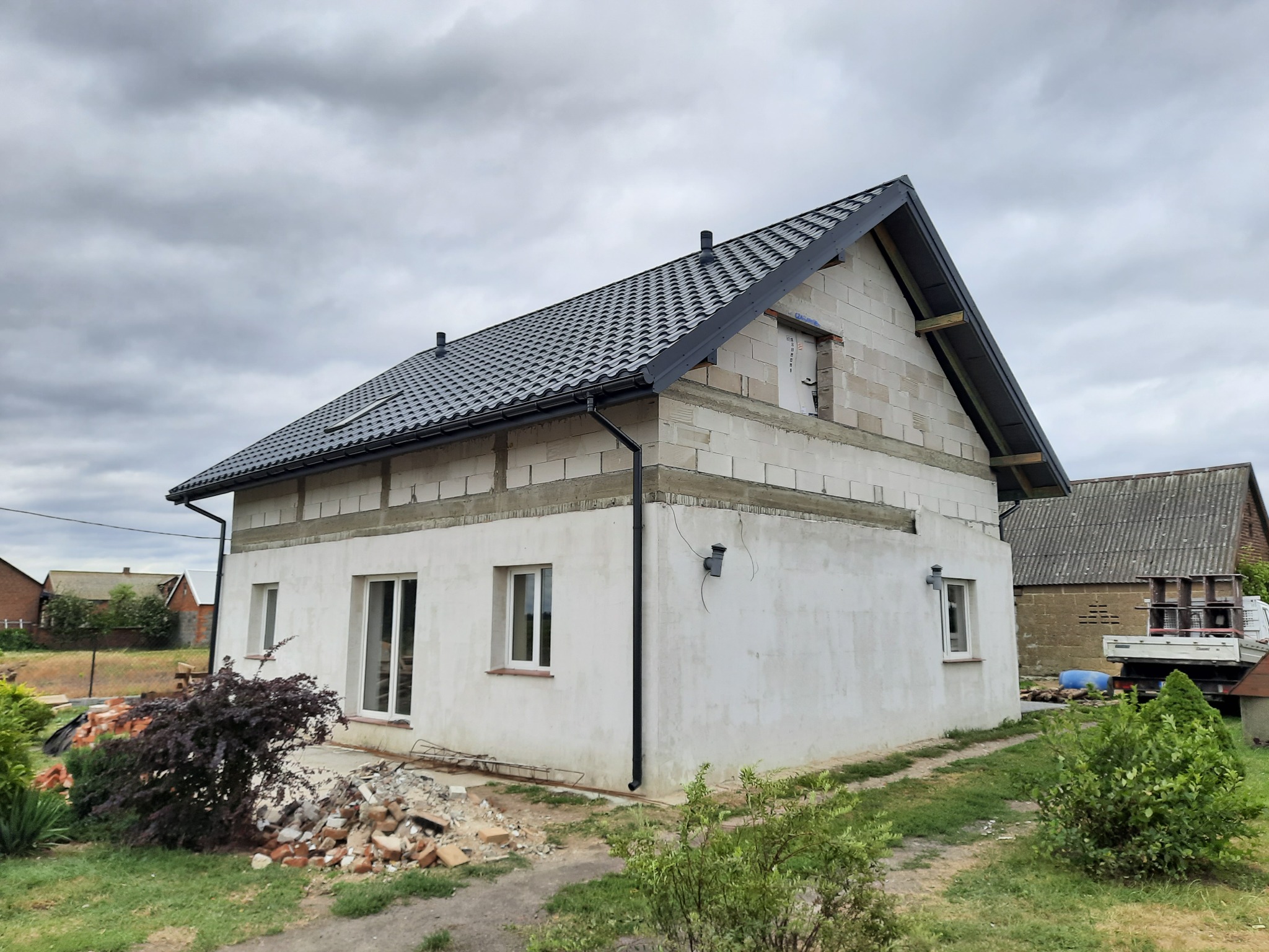 Surowy stan domu jednorodzinnego z szarą dachówką, widoczne bloczki Ytong i częściowo położony tynk zewnętrzny, gruz budowlany przed budynkiem.