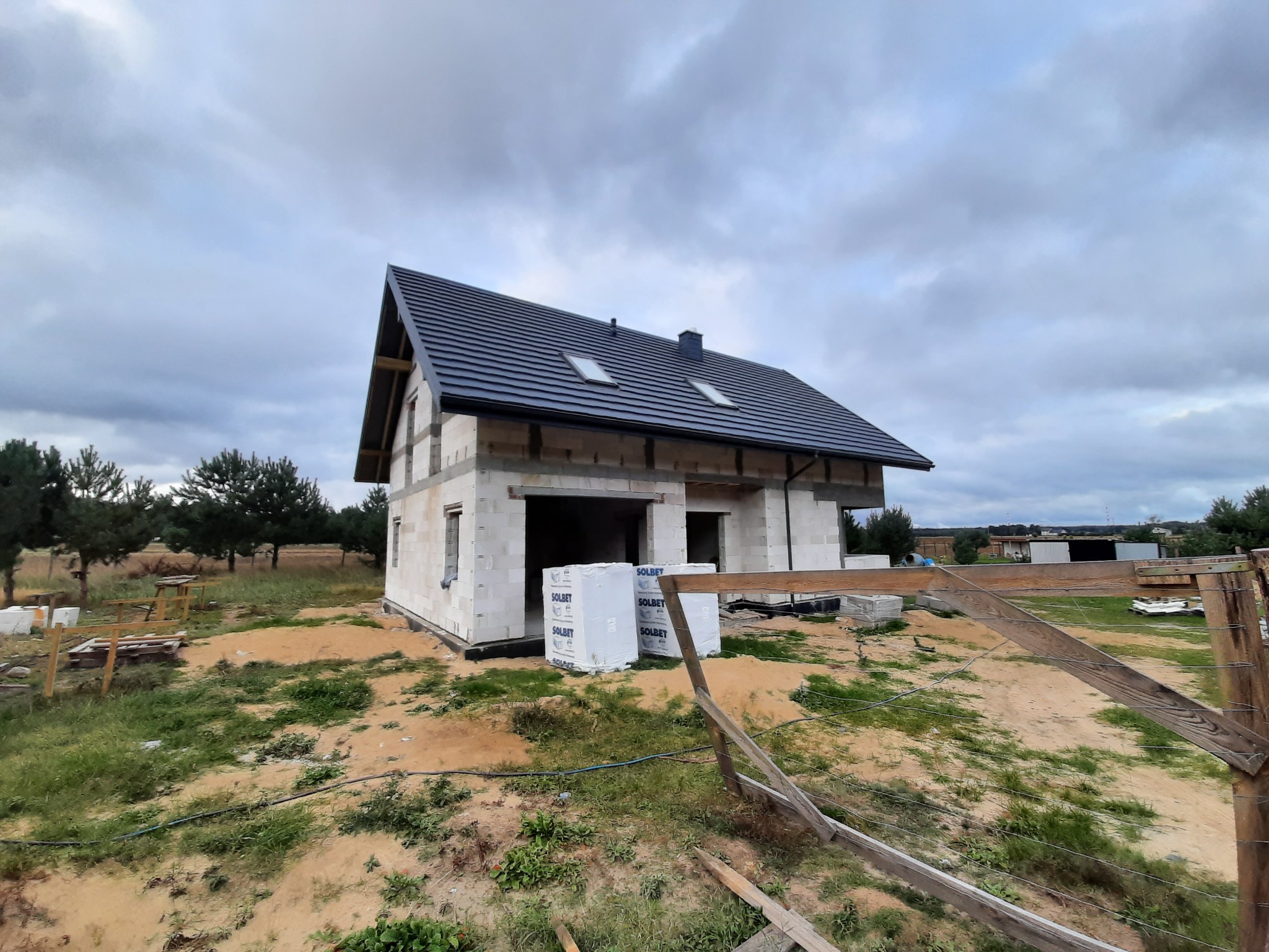 Surowy stan budowy domu z białych bloczków Solbet, z widocznym dachem z ciemnej dachówki i oknami dachowymi, na działce z elementami drewnianego ogrodzenia i stertą materiałów budowlanych.