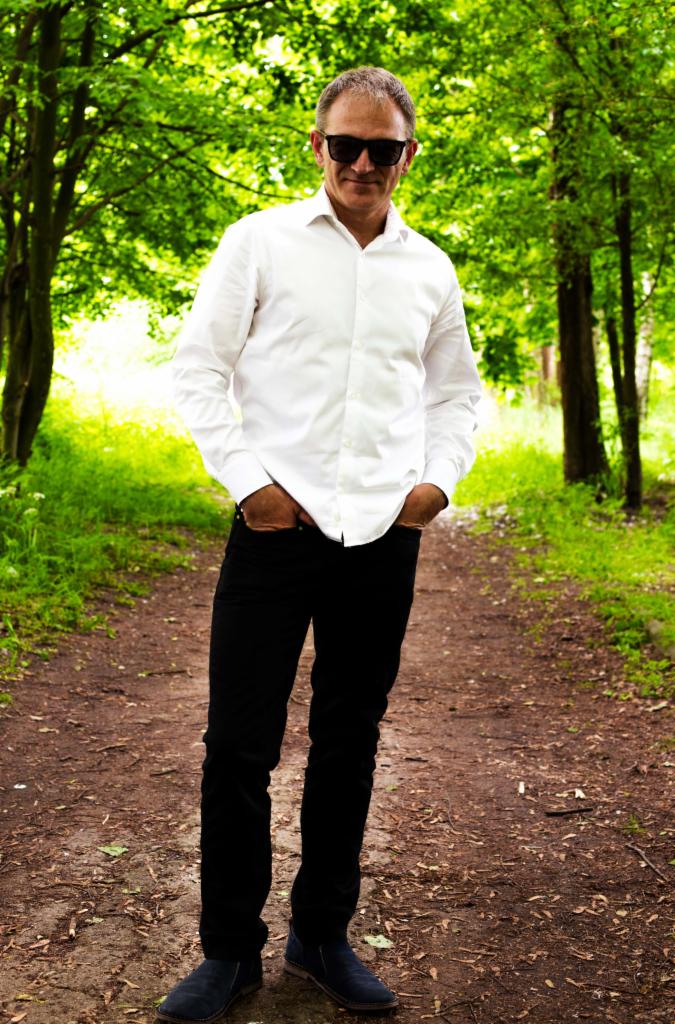 Mężczyzna w białej koszuli i czarnych spodniach, z rękami w kieszeniach, stoi na leśnej ścieżce w Pabianicach. Ubrany w ciemne okulary, pozuje na tle zieleni.