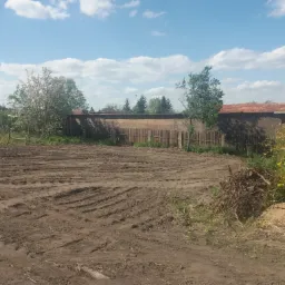 Przygotowany teren pod budowę na działce, widoczne ślady po maszynach, w tle ogrodzenie i budynki.