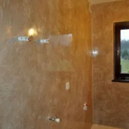 Wykończona łazienka z beżowymi ścianami o marmurkowym wzorze, widoczne przyłącza wodne i okno z widokiem na zieleń.