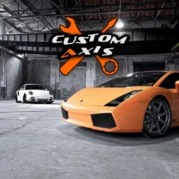 Pomarańczowy Lamborghini Gallardo i białe Porsche 911 w przestronnym warsztacie z betonową podłogą i ścianami z cegieł. W tle widoczne logo warsztatu 'Custom Axis' z kluczami.