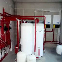 Kotłownia z dwoma zbiornikami buforowymi, czerwonymi rurami grzewczymi z izolacją, dwoma białymi kotłami grzewczymi na ścianie oraz białymi rurami instalacji elektrycznej pod sufitem.