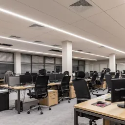 Nowoczesne oświetlenie LED w sali biurowej, liniowe oprawy sufitowe, biurka z komputerami i krzesłami biurowymi, widok na okna z żaluzjami.