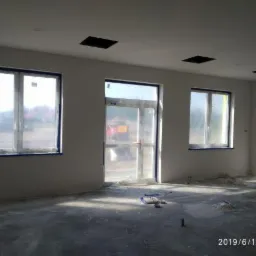 Wnętrze pomieszczenia w trakcie remontu: ściany przygotowane do prac wykończeniowych, okna zabezpieczone folią, widoczne ślady po szpachlowaniu na podłodze.