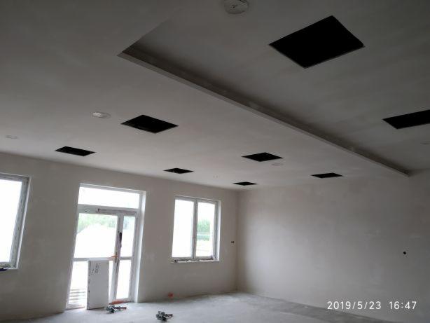 Wnętrze w trakcie szpachlowania ścian i sufitu z przygotowanymi otworami na oświetlenie, widoczne ślady po szpachli na ścianach i suficie, okna z widokiem na zewnątrz, narzędzia na podłodze.