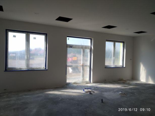 Wnętrze pomieszczenia w trakcie remontu: ściany przygotowane do prac wykończeniowych, okna zabezpieczone folią, widoczne ślady po szpachlowaniu na podłodze.