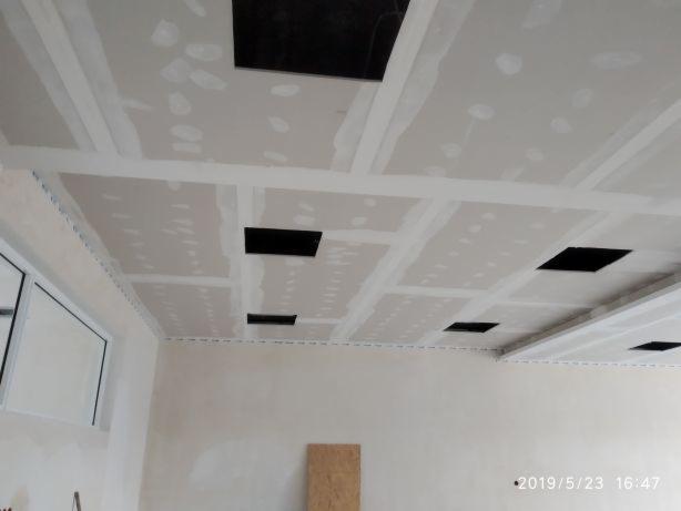 Sufit z płyt kartonowo-gipsowych w trakcie montażu, widoczne otwory na oświetlenie, łączenia wzmocnione taśmą, ściany w tle przygotowane do dalszych prac wykończeniowych.