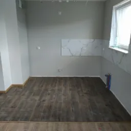 Wnętrze pomieszczenia po remoncie: położona drewnopodobna podłoga, fragment ściany z płytkami imitującymi marmur, widoczne rury instalacji wodnej przy ścianie pod oknem.
