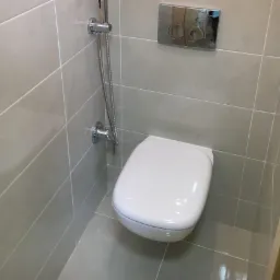 Nowoczesna toaleta podwieszana z chromowaną armaturą bidetową i przyciskiem spłukującym na szarych, ceramicznych płytkach.