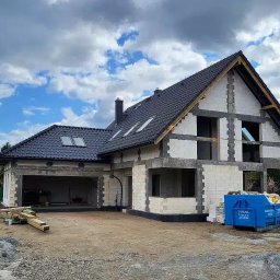 Solidbud - Surowy stan domu murowanego z garażem w trakcie budowy, widoczne bloczki, dach z dachówką, okna bez szyb, kontener na gruz. Niebo pochmurne.