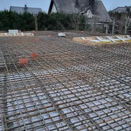 Solidbud - Zbrojenie stropu budynku jednorodzinnego z widocznymi elementami szalunku i dystansami z tworzywa. Siatka z prętów stalowych na tle zabudowy mieszkalnej.