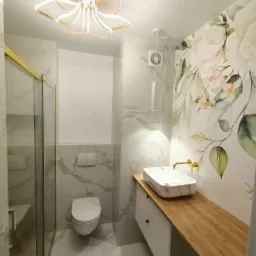 Nowoczesna łazienka z marmurowymi płytkami, umywalką nablatową z drewnianym blatem, kabiną prysznicową ze złotymi elementami oraz dekoracyjną tapetą w kwiaty.