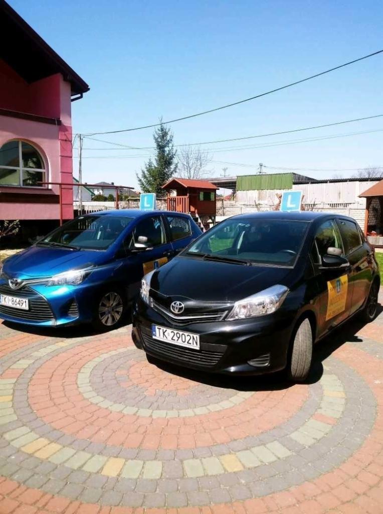 Dwa samochody Toyota Yaris z literą 'L' na dachu, zaparkowane na brukowanym podjeździe przed budynkiem.