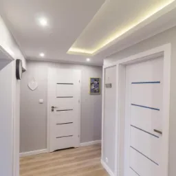 Nowoczesny korytarz z dwiema białymi drzwiami z poziomymi paskami, drewnopodobną podłogą, podwieszanym sufitem z oświetleniem LED i szarą ścianą z dekoracjami.