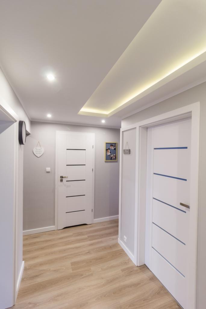 Nowoczesny korytarz z dwiema białymi drzwiami z poziomymi paskami, drewnopodobną podłogą, podwieszanym sufitem z oświetleniem LED i szarą ścianą z dekoracjami.