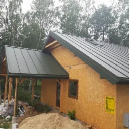 Surowy stan domu szkieletowego z zamontowanym, grafitowym dachem z blachy trapezowej i oknem dachowym, widoczne drewniane elementy konstrukcji i sterta piasku przy ścianie.