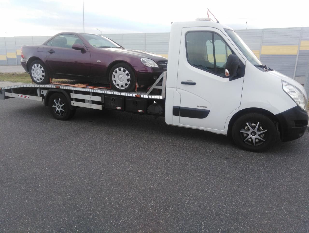 Bordowy Mercedes SLK na białej lawecie na tle szarego ekranu akustycznego.