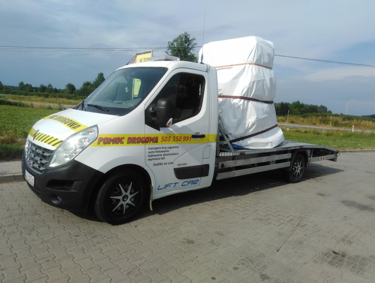 Biała laweta z napisem 'Pomoc Drogowa' przewozi zabezpieczony ładunek. Widoczne numery kontaktowe i informacje o usługach. Tło stanowi zielony krajobraz.