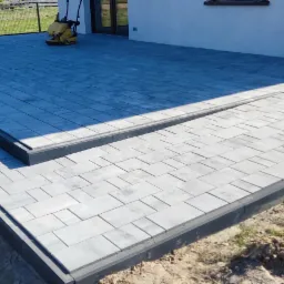 Nowoczesny taras z szarej kostki brukowej, z dwoma stopniami i obrzeżami, widok na ogród w tle, częściowo zacieniony.