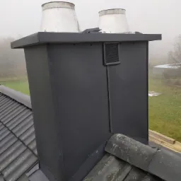 Czarny komin z dwoma białymi wylotami wentylacyjnymi na ciemnym, ceramicznym dachu w pochmurny dzień, widoczny fragment ogrodu w tle.