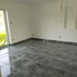 Wykończone pomieszczenie z szarą, marmurkową podłogą z płytek, białe ściany z przygotowanymi punktami elektrycznymi, widok przez okno na zieleń.