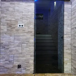 Szklane drzwi przesuwne z czarną prowadnicą, prowadzące do schodów, na tle ściany wyłożonej płytkami imitującymi kamień i marmurowej podłogi.