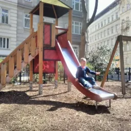 Chłopiec zjeżdża ze zjeżdżalni na placu zabaw, w tle budynki mieszkalne i drzewa.