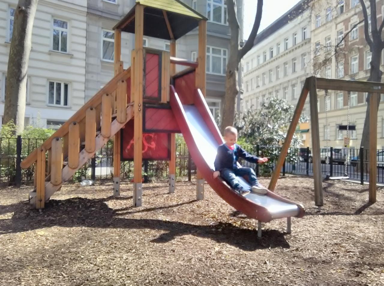 Chłopiec zjeżdża ze zjeżdżalni na placu zabaw, w tle budynki mieszkalne i drzewa.