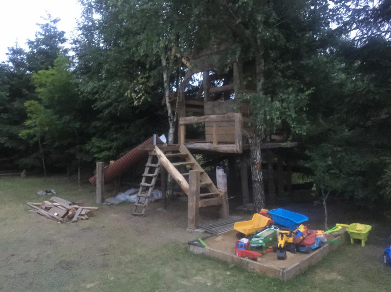 Drewniany plac zabaw na podwórku z domkiem na drzewie, zjeżdżalnią z rury, schodami i piaskownicą wypełnioną zabawkami.