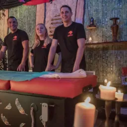 Trzy osoby w czarnych koszulkach z logo firmy stoją za czerwonym łóżkiem do masażu w przytulnym, oświetlonym świecami pomieszczeniu z drewnianymi elementami dekoracyjnymi i piórami ozdobnymi na łóżku.