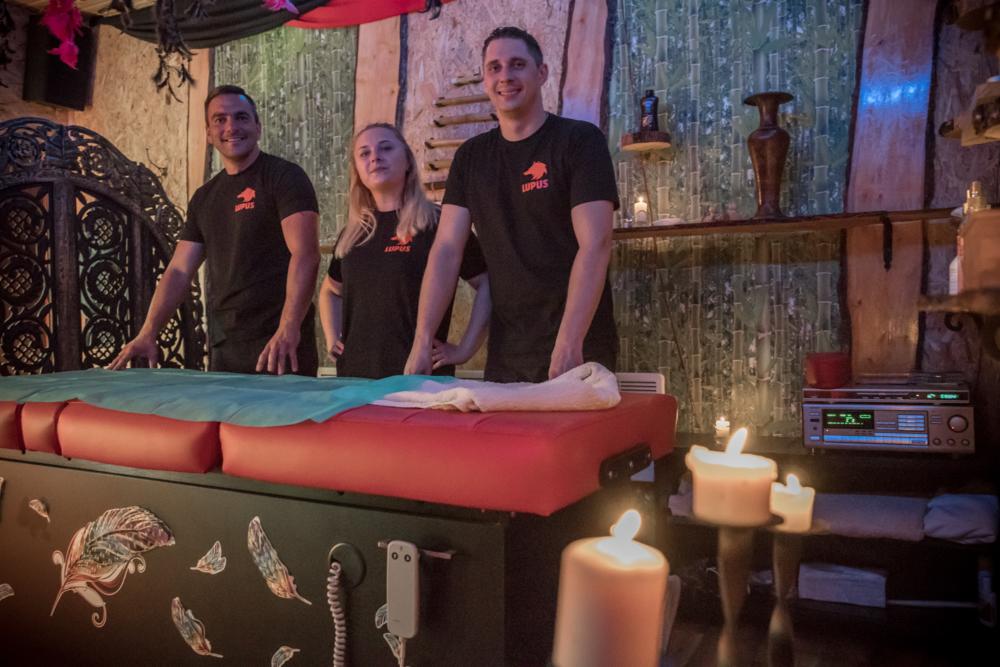 Trzy osoby w czarnych koszulkach z logo firmy stoją za czerwonym łóżkiem do masażu w przytulnym, oświetlonym świecami pomieszczeniu z drewnianymi elementami dekoracyjnymi i piórami ozdobnymi na łóżku.