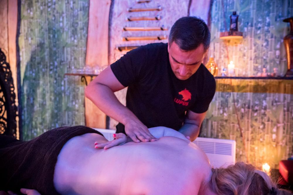 Mężczyzna w czarnej koszulce z logo Lupus wykonuje masaż pleców klientki w klimatycznym pomieszczeniu z dekoracjami imitującymi naturalne środowisko, przy stonowanym oświetleniu świec.