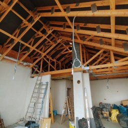 TB wykończenia wnętrz Tomasz Bargieła - Konstrukcja drewniana dachu z izolacją, widoczne elementy instalacji elektrycznej i wentylacyjnej. Dwie drabiny i materiały budowlane w surowym wnętrzu budynku.