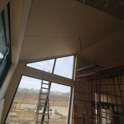 TB wykończenia wnętrz Tomasz Bargieła - Skośny sufit z płyt karton-gips w nowoczesnym domu z dużymi oknami. Widoczne rusztowania i drabina, co sugeruje trwający montaż. Nietypowy kształt okien.