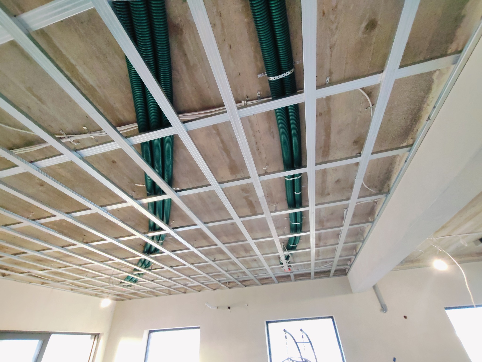 Konstrukcja sufitu podwieszanego z widocznymi zielonymi rurami instalacyjnymi przymocowanymi do stropu za pomocą metalowych obejm.