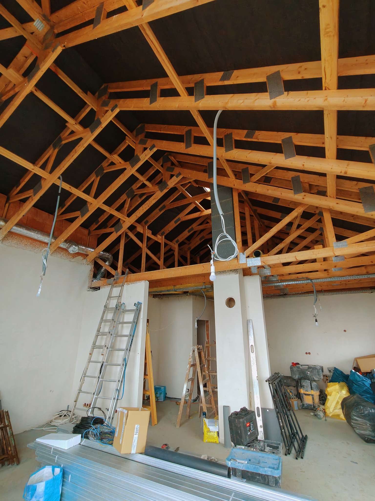 Konstrukcja drewniana dachu z izolacją, widoczne elementy instalacji elektrycznej i wentylacyjnej. Dwie drabiny i materiały budowlane w surowym wnętrzu budynku.