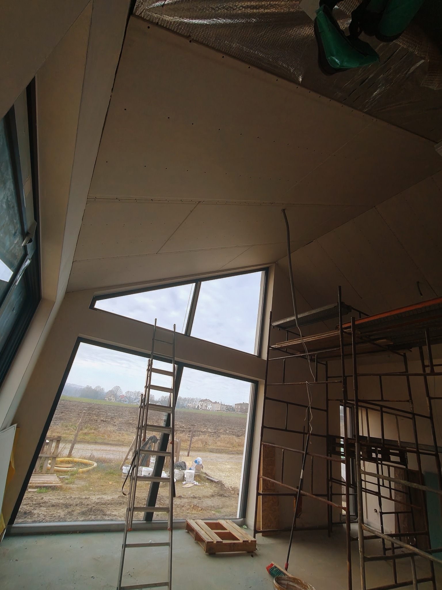 Skośny sufit z płyt karton-gips w nowoczesnym domu z dużymi oknami. Widoczne rusztowania i drabina, co sugeruje trwający montaż. Nietypowy kształt okien.