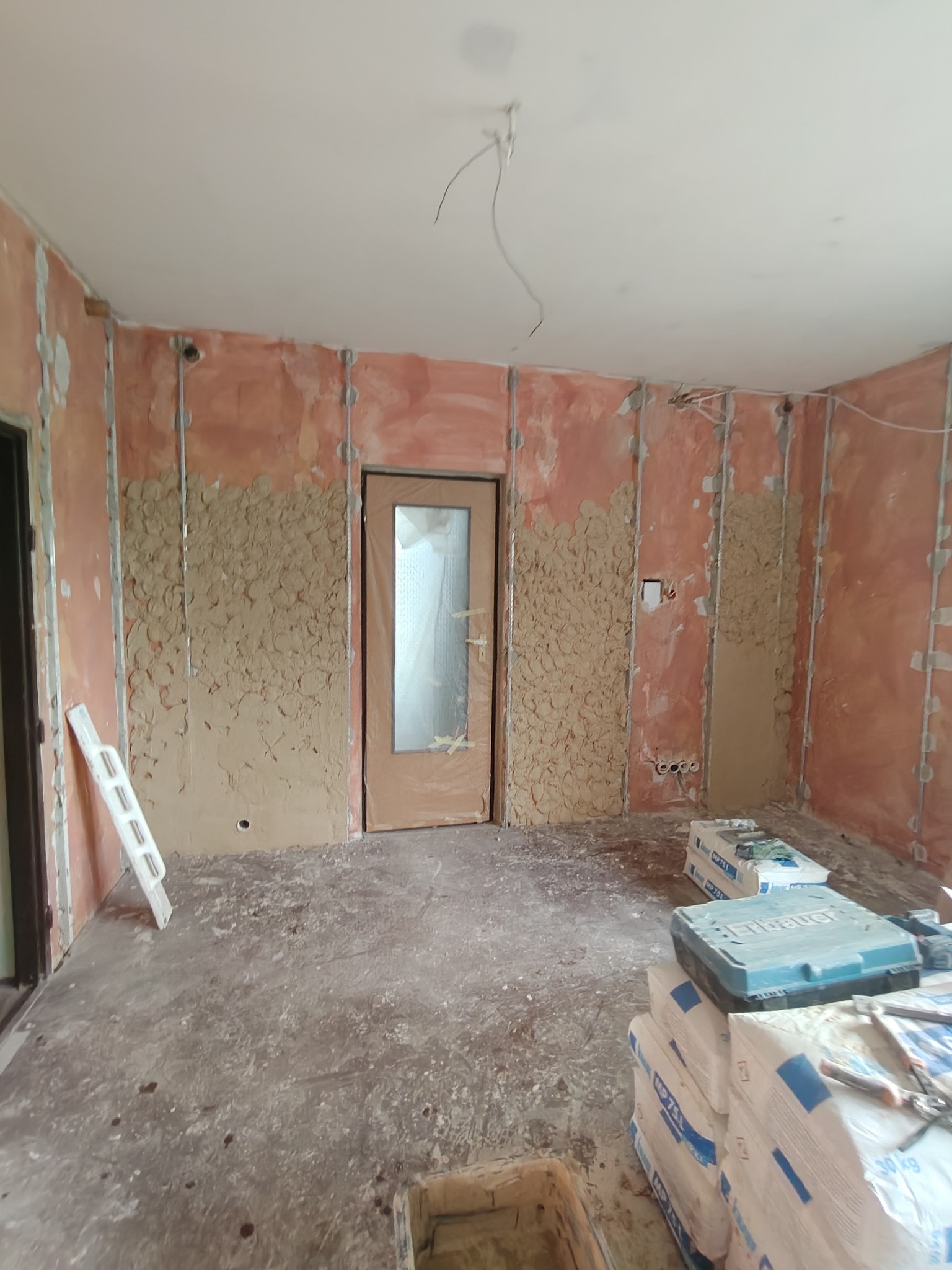 Wnętrze w trakcie prac wykończeniowych: ściany z nałożoną warstwą wstępną gładzi, widoczne profile, worki z zaprawą i narzędzia. Drzwi zabezpieczone folią.