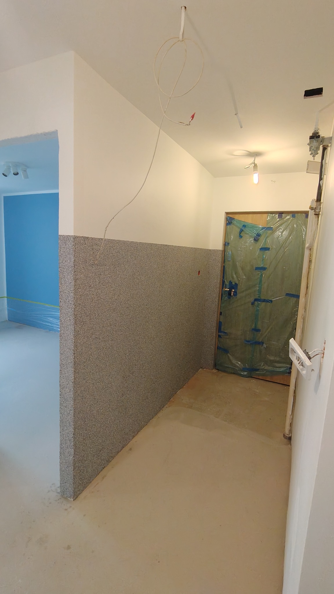 Korytarz w trakcie prac wykończeniowych: ściana z szorstką fakturą do połowy wysokości, zabezpieczone folią drzwi, widoczne przewody elektryczne.