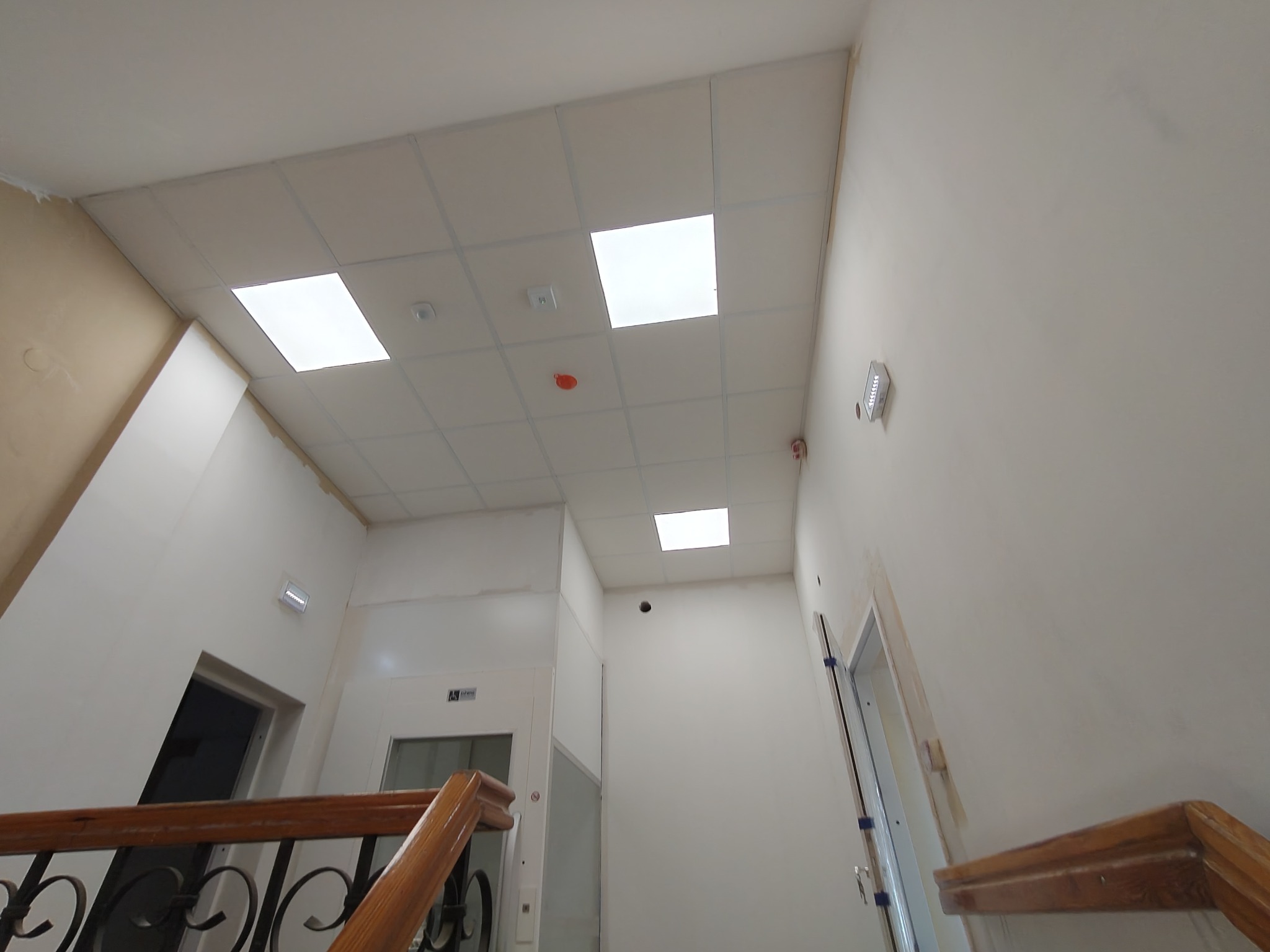 Klatka schodowa po remoncie: białe ściany, sufit kasetonowy z oświetleniem, drewniana balustrada. Widoczne ślady po pracach wykończeniowych.