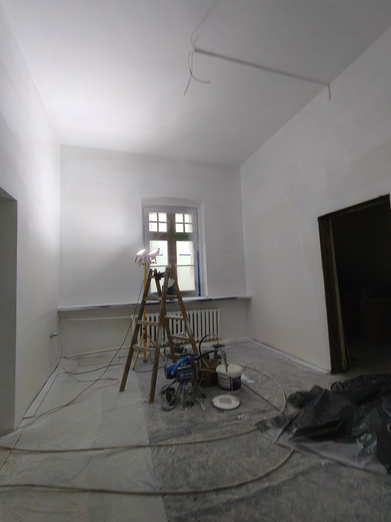 Białe ściany po malowaniu natryskowym w pokoju z oknem, drabina, sprzęt malarski i zabezpieczona podłoga. Widok na odnowione wnętrze.