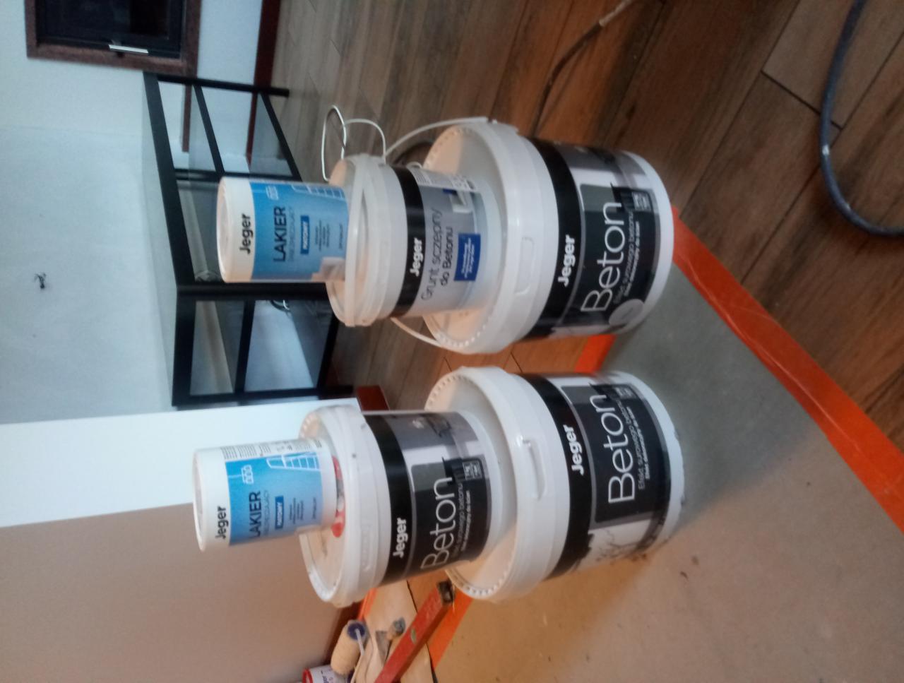 Dwa stosy białych wiader z farbą i gruntem marki Jeger, w tym wiadro z napisem 'Beton', ustawione na zabezpieczonej folią podłodze podczas prac malarskich.