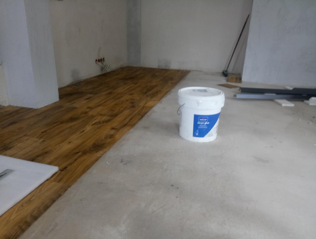 Fragment układania paneli podłogowych w pomieszczeniu, widoczny biały pojemnik z klejem i częściowo ułożona podłoga o drewnopodobnym wzorze.